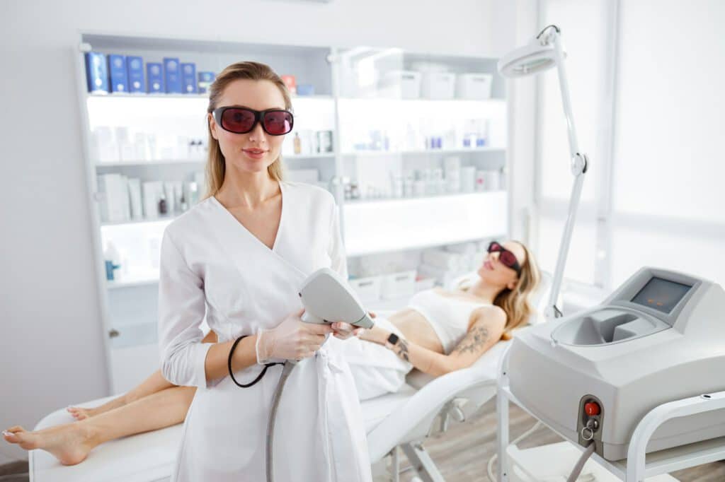 Beautician with laser device and woman on daybed in beauty salon